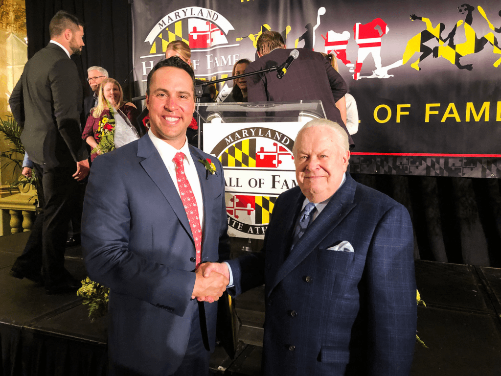 Mark Teixeira shaking hands with an older man at an event, both smiling, with a Maryland State Athletic Hall of Fame backdrop in the background.