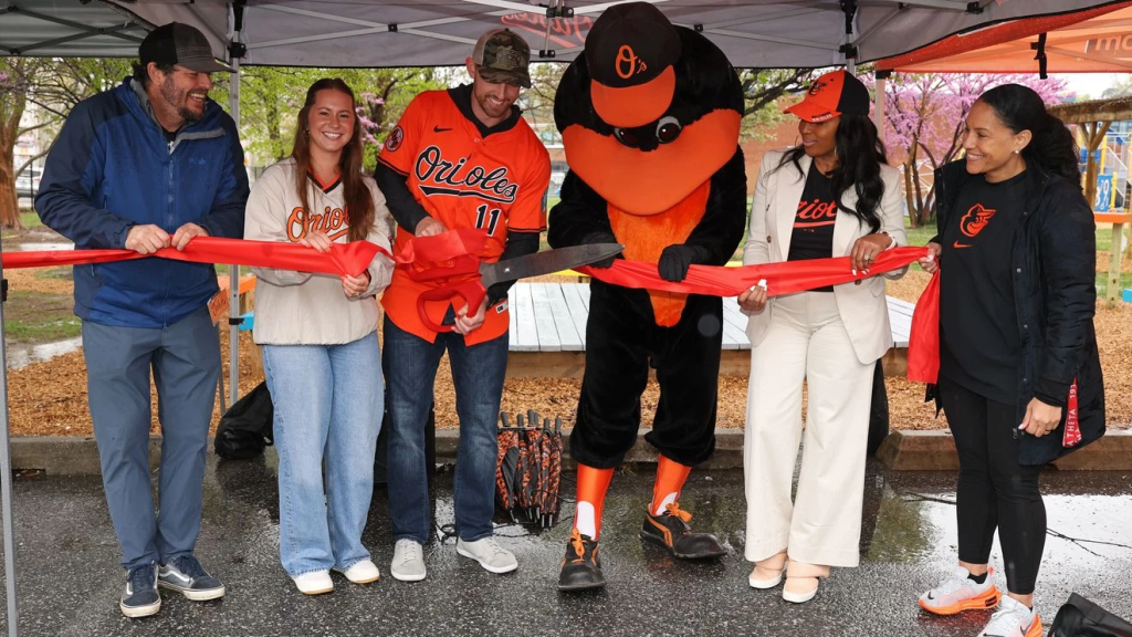 A group of five people, including a mascot, stand together under a tent, holding a red ribbon for a ceremonial ribbon-cutting event. They are wearing Baltimore Orioles apparel, and the scene appears to be set outdoors on a rainy day.