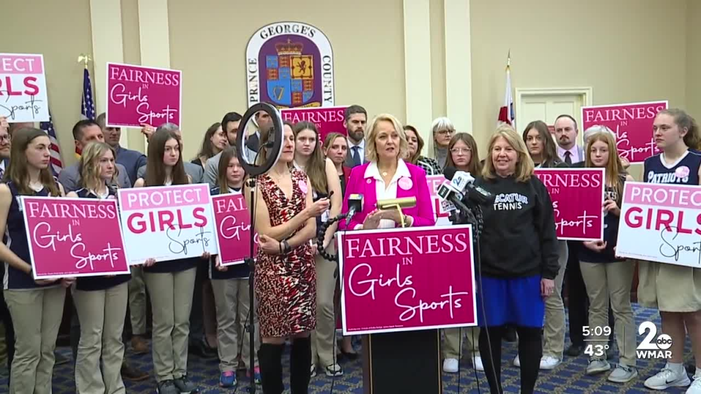Group of individuals, including girls and adults, holding signs that promote fairness in girls' sports during a press conference.